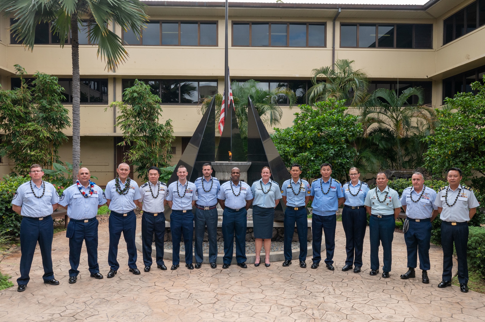 A group of service members stand in a horizontal line in front of the memorial in the Courtyard of Heroes, where an eternal flame is lit and flickering behind them.