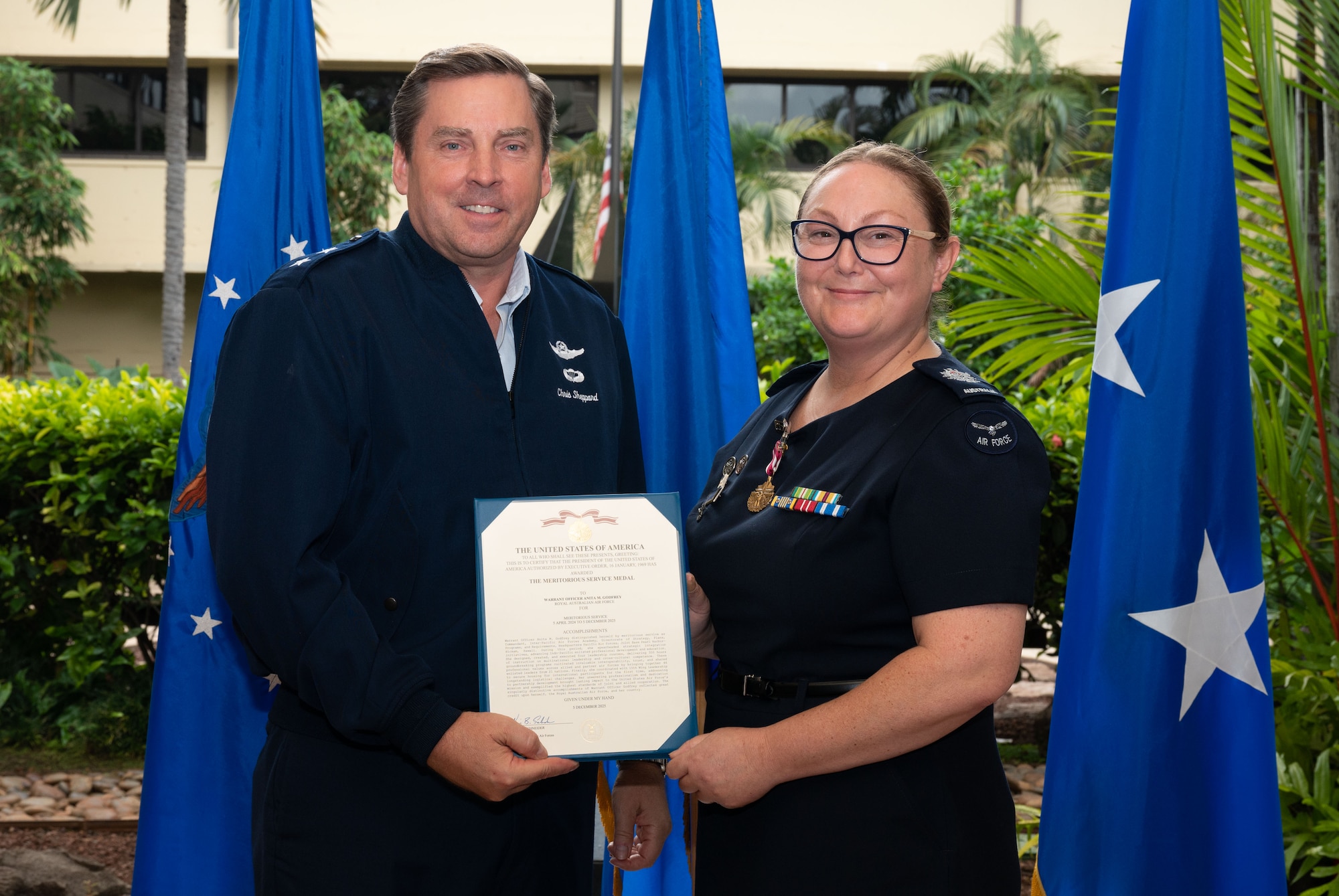 A male U.S. major general to the left and female Royal Australian Air Force warrant officer, on the right, wearing a meritorious service medal on her chest, stand together for a photo holding the meritorious service award memorandum.