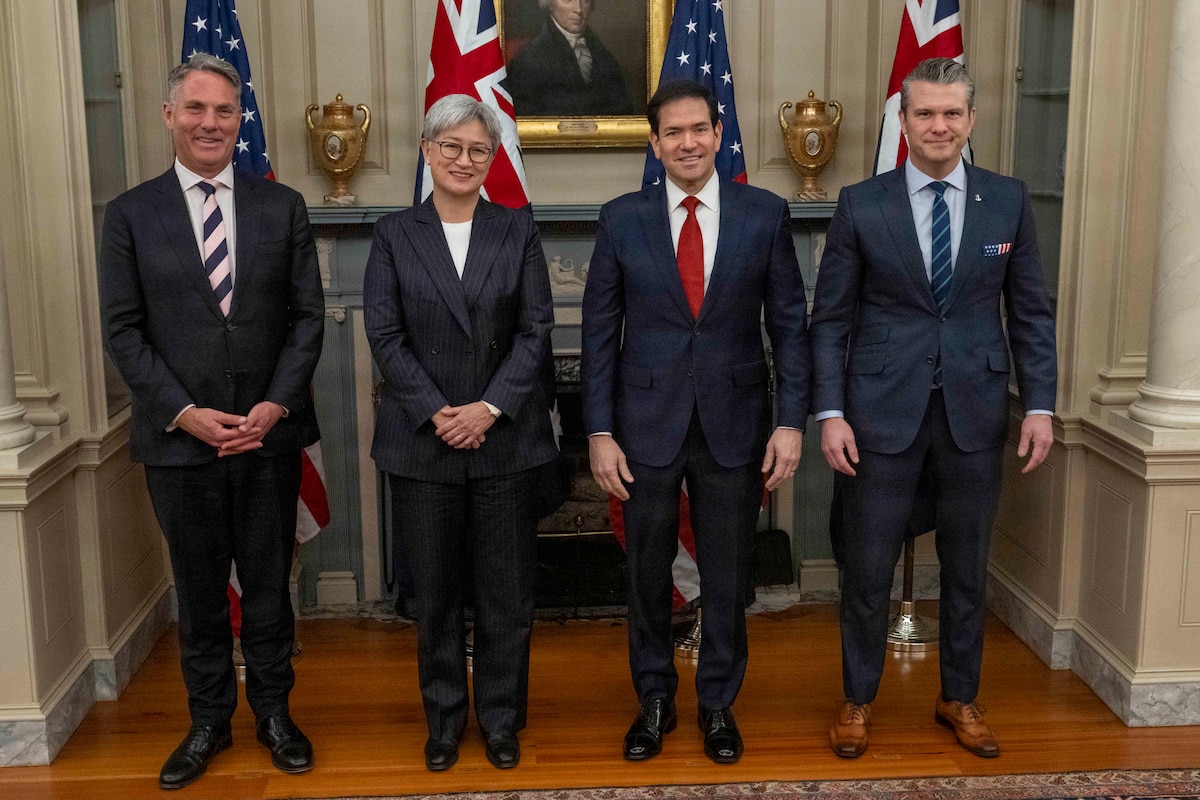 Three men and one woman, all wearing business attire, stand together, with alternating American and Austrian flags behind them.