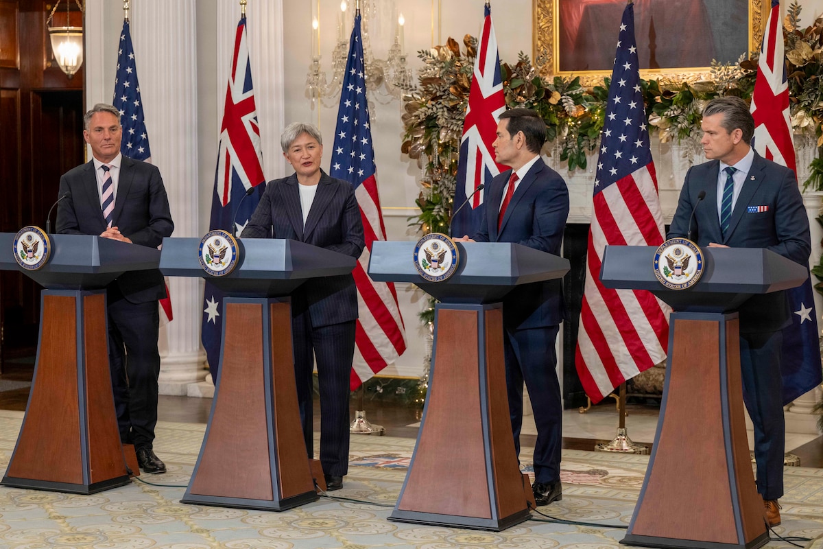 Three men and one woman, all wearing business attire, stand at lecterns with alternating American and Austrian flags behind them.