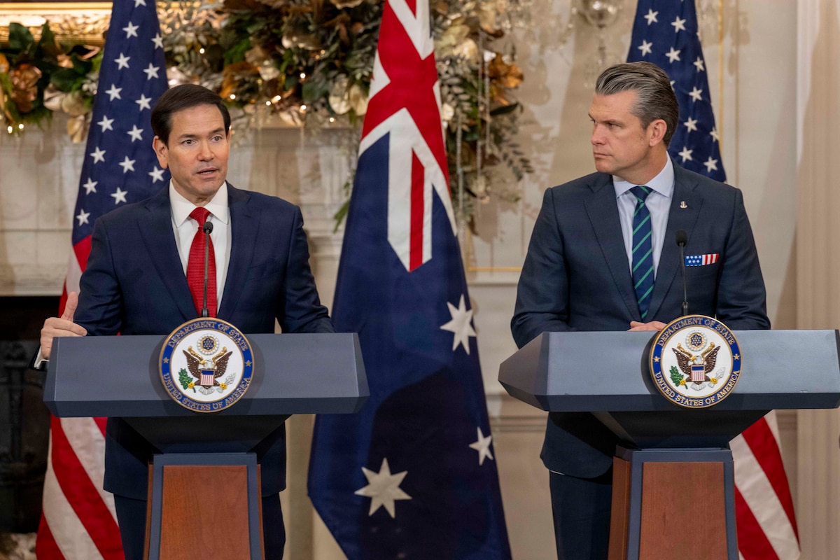 A man wearing business attire stands at a lectern and speaks as another man in similar attire looks on; behind them are alternating American and Austrian flags.