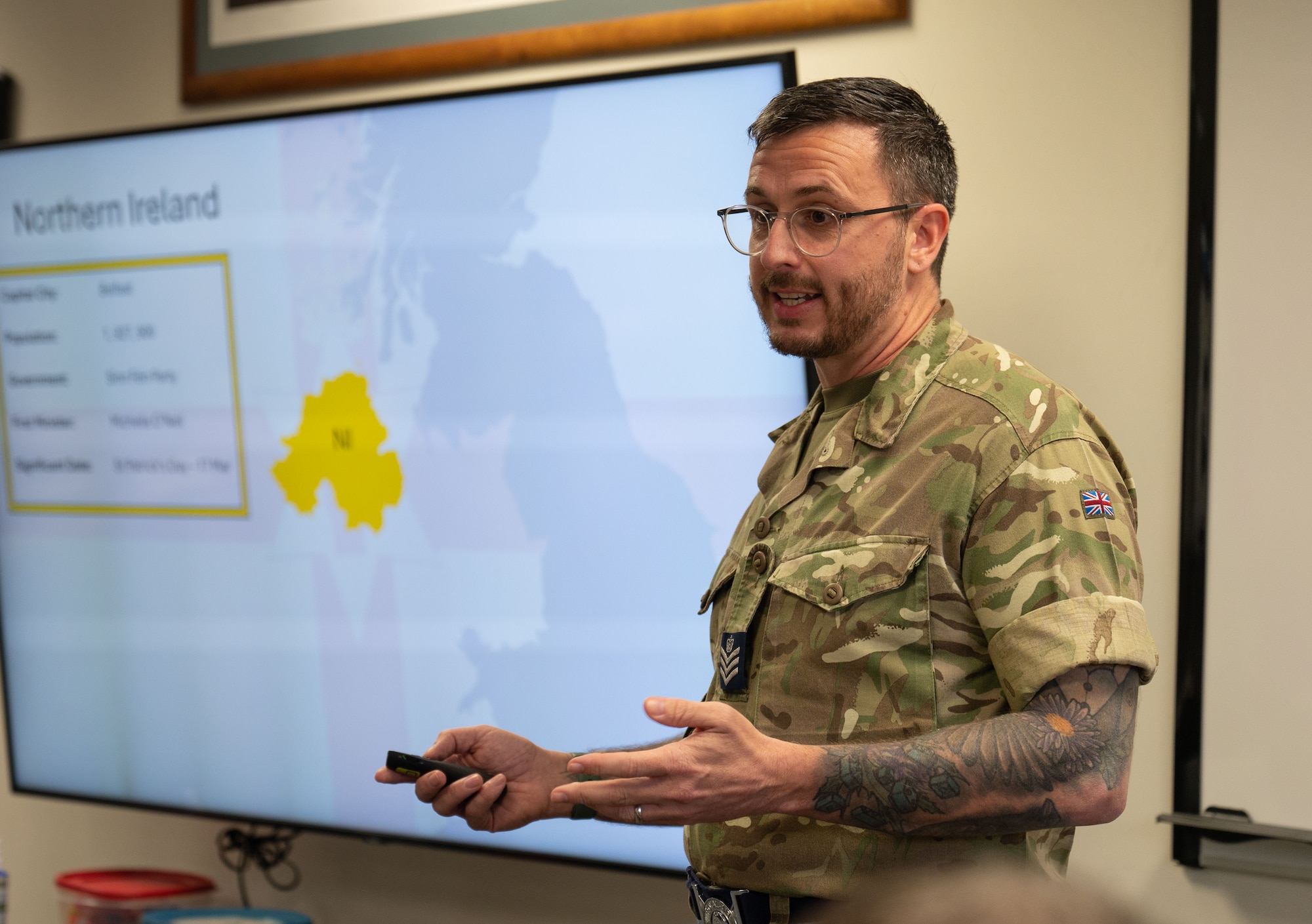 A close up of a male Royal Air Force military member speaking in front of a presentation with a map.