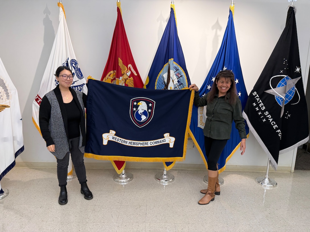two women hold up a blue flag with an eagle, globe and written scroll, with gold fringe