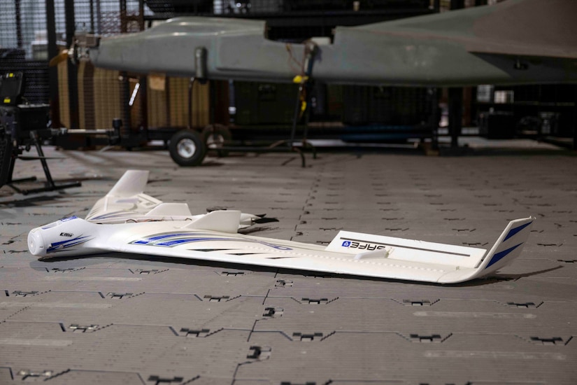 A small, white drone sits on the floor. A larger drone sits on wheels in the background.