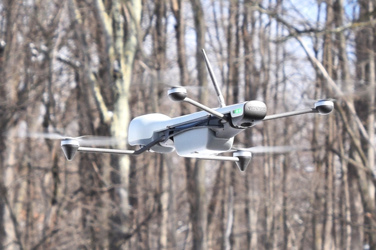 A quadcopter drone flies in the air amid a backdrop of leafless trees.