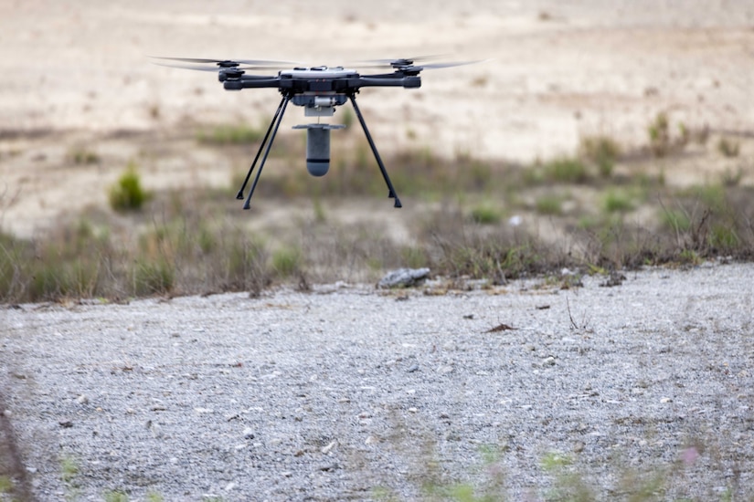 A quadcopter drone hovers a few feet above gravel.