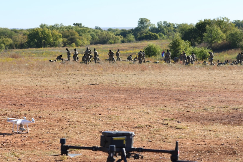 Two drones sit on a dirt patch. In the distance, several soldiers mill around.