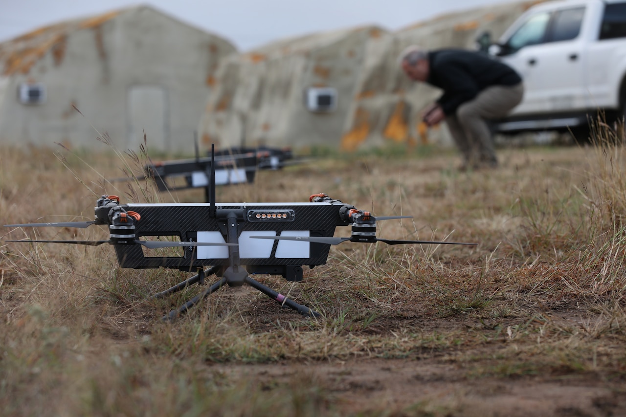 A small quadcopter drone sits on grass. In the background, another drone sits, and a man bends over to look at it.