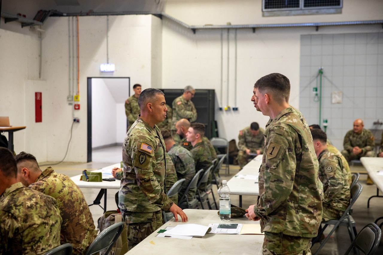 A soldier in a camouflage military uniform stands in front of a table while talking to two soldiers in similar attire standing across from him. Several other people in similar attire are seated at tables around the room.