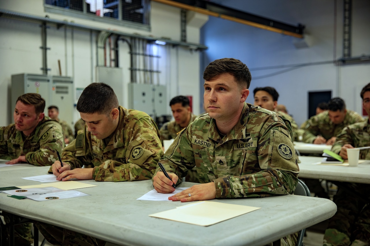 Three soldiers in camouflage military uniforms write on pieces of paper while seated at a table. Several other soldiers in similar attire, doing the same thing, are seated at a table in the background.