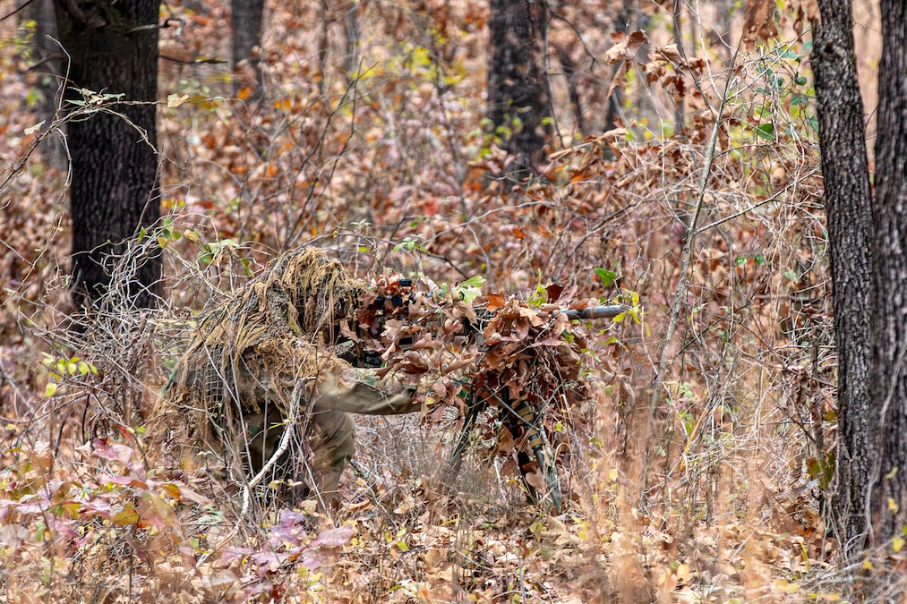 A competitor hides among leaves and branches while aiming a sniper rifle in the woods.