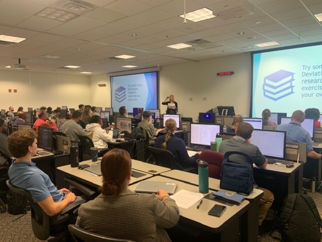 A classroom full of computer stations and students sitting at them.