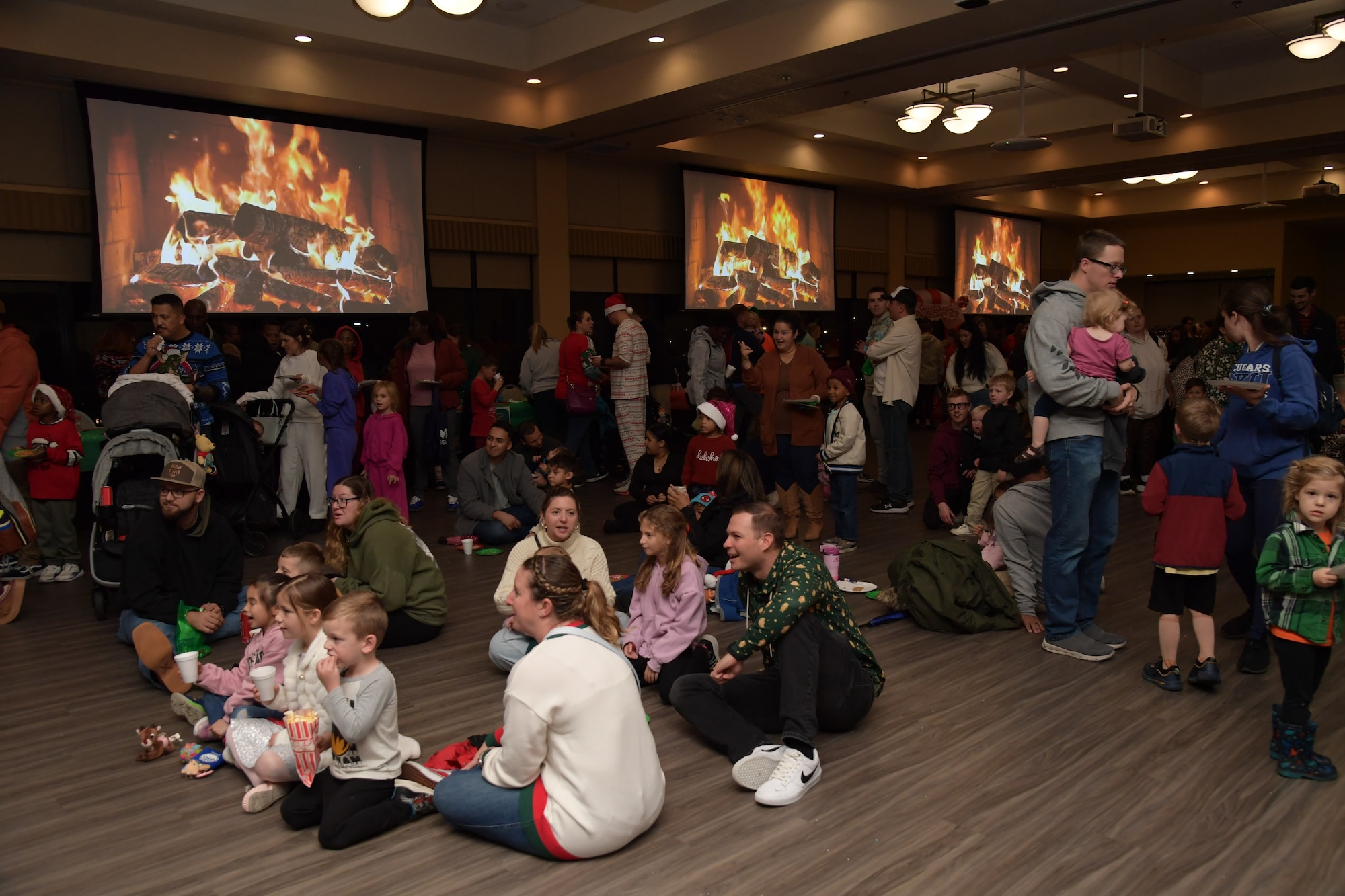 A group of people sit and stand around a room with monitors displaying a fireplace hanging on the wall.