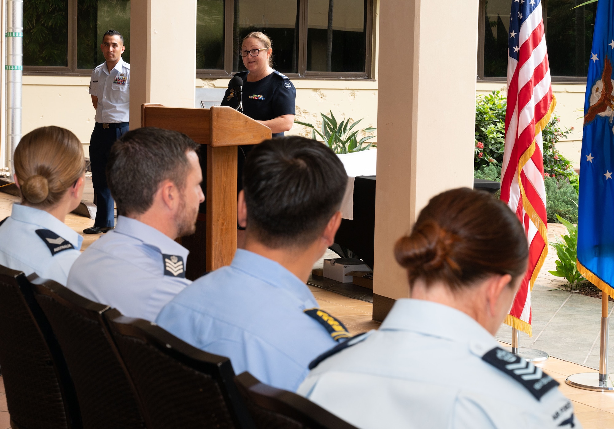 A female Royal Australian Air Force Warrant Officer stands at a podium speaking diagonally from four graduates of different national militaries seated.