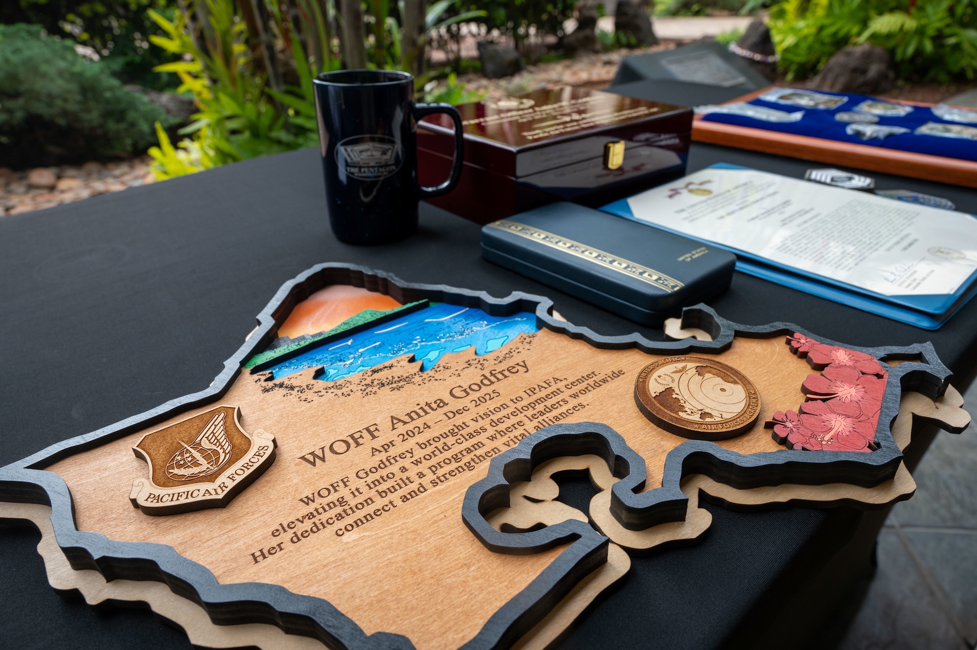 On a table a Pacific Air Forces wood plaque of appreciation is in focus. It is in the shape of the state of Hawaii and has a raised border and raised design elements of 3 Hibiscus flowers, Hawaii's state flower, and a coin shape engraved map of the Indo-Pacific with a point of signal and bordered with stars at the end of "Inter-Pacific" along the top of the coin shape and "Air Forces Academy" along the bottom. 

There is also a cutout of the ocean with specs of sand surrounding it in the top corner of the state shape. Another raised design on the wooden plaque is the Pacific Air Forces shield engraved with it's symbol of a globe with a lightning bolt in it, a sprig lying beside the lightning bolt still in the globe and four stars in the globe above the sprig next to the lightning bolt. Coming off the globe's right are wings and the bottom of the shield reads "Pacific Air Forces". 

In the center of the plaque reads: 
"WOFF Anita Godfrey
Apr 2024 - Dec 2025
WOFF Godfrey brought vision to IPAFA, elevating it into a world-class development center. Her dedication built a program where leaders worldwide connect and strengthen vital alliances."