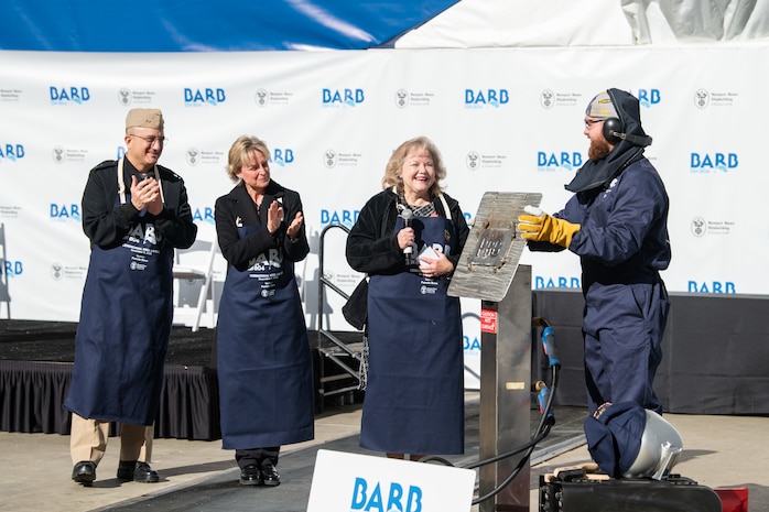 From left, Rear Adm. Casey Moton, Program Executive Officer, Aircraft Carriers, Newport News Shipbuilding President Kari Wilkinson, and ship’s sponsor Pam Bove, applaud NNS welder Andrew Kahler after he welded the initials of Bove onto a metal plate during the keel authentication ceremony for Virginia-class attack submarine Barb (SSN 804) on Tuesday, Dec. 9, 2025 at HII’s Newport News Shipbuilding division. The metal plate will remain affixed to the submarine throughout its life.
