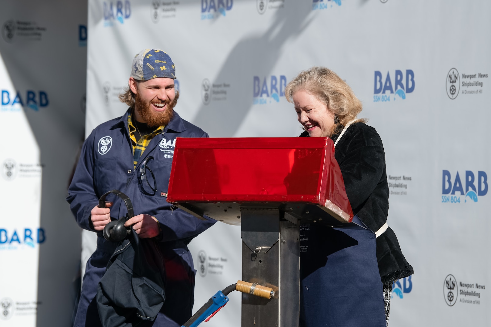 At the weld stand, ship’s sponsor Pam Bove traces her initials on a metal plate during the keel laying ceremony for Virginia-class attack submarine Barb (SSN 804) at HII’s Newport News Shipbuilding division on Tuesday, Dec. 9, 2025. At left is NNS welder Andrew Kahler.
