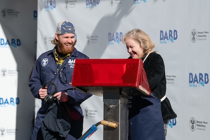 At the weld stand, ship’s sponsor Pam Bove traces her initials on a metal plate during the keel laying ceremony for Virginia-class attack submarine Barb (SSN 804) at HII’s Newport News Shipbuilding division on Tuesday, Dec. 9, 2025. At left is NNS welder Andrew Kahler.