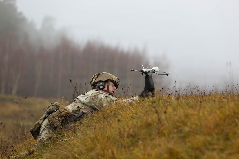 A soldier wearing a camouflage military uniform lies in a grassy field and prepares to launch a small drone on a cloudy day.