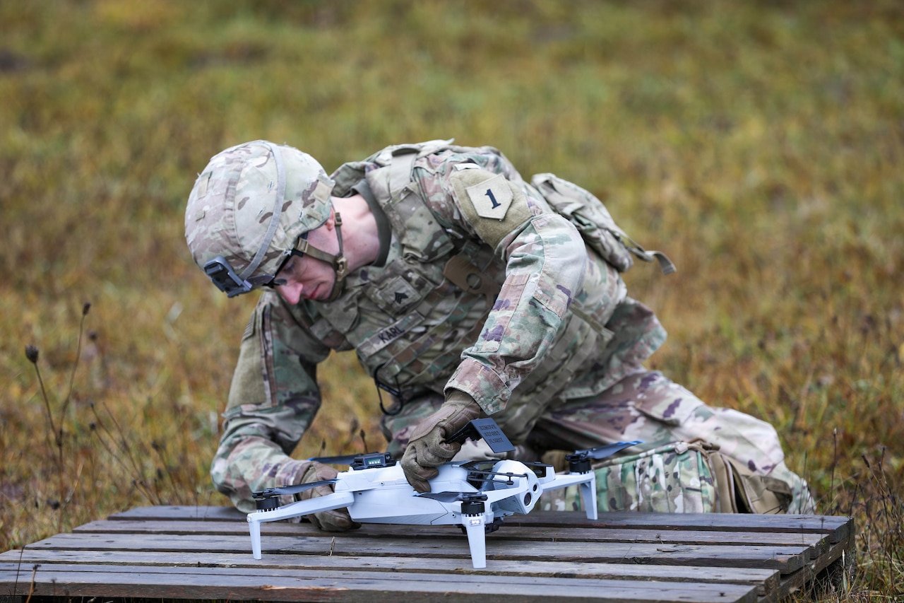 A soldier wearing a camouflage military uniform steadies a small drone on a wooden table outside and prepares it for launch.