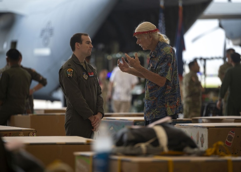 Two men, one a service member and the other a civilian, chat amongst a sea of supply boxes in an aircraft hangar. A military aircraft is in the background.