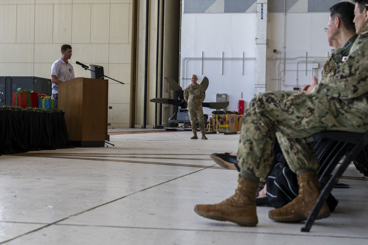 A man stands at a lectern and gives a speech to a seated crowd of civilians and service members inside an aircraft hangar.