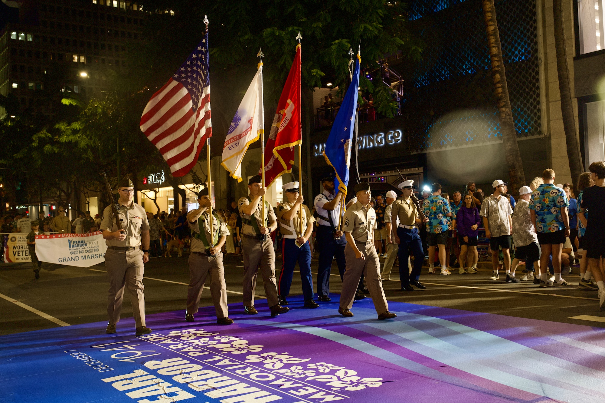 A joint service color guard, consisting of five men led by another man, all in military dress per their branch of service, walks in a parade holding flags.