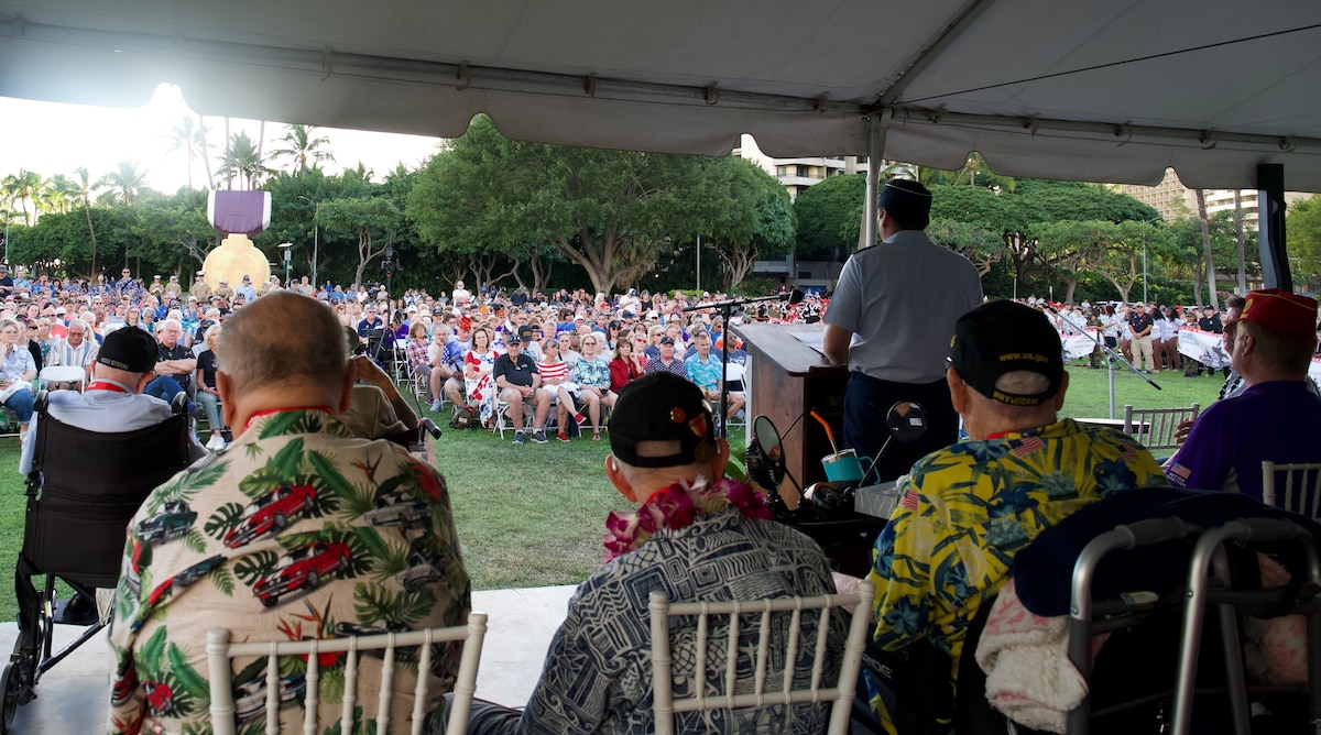 A male senior military officer gives a speech to a crowd of hundreds from a podium on a stage.