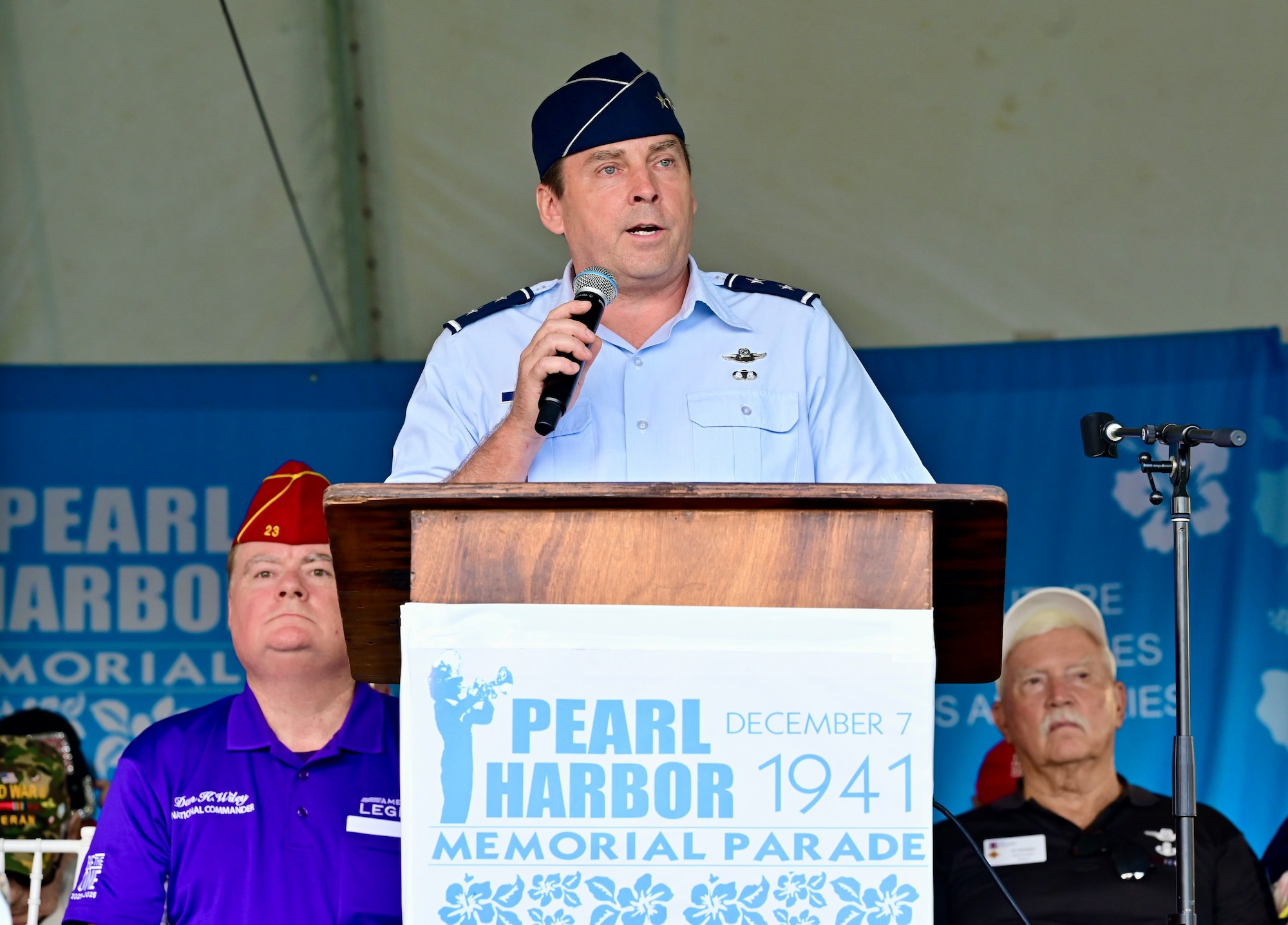 A male senior officer speaks into a microphone at a podium on stage. The podium has a sign read: "Pearl Harbor Memorial Parade December 7 1941."
