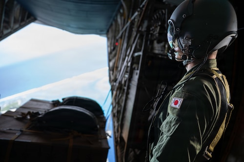 A person with a helmet looking over an open ramp of an aircraft mid-flight