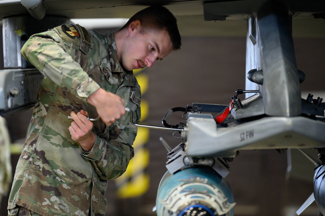 A person uses a tool underneath the wing of a fighter aircraft to secure two bombs.