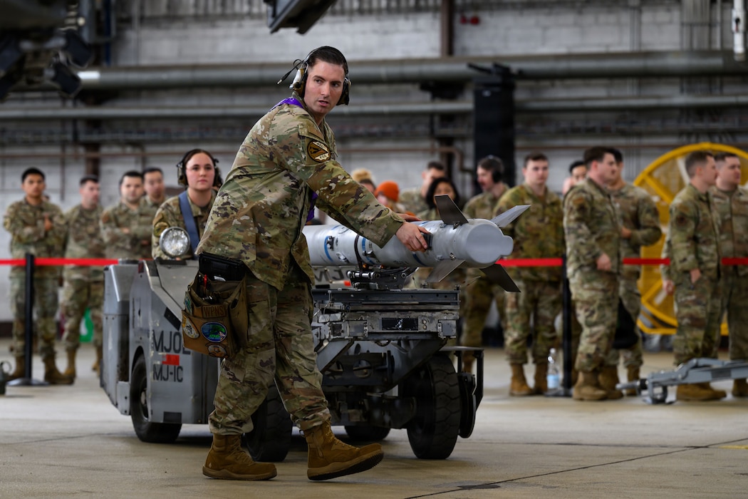 A team of people help transport a weapon on a loading vehicle towards a fighter aircraft.