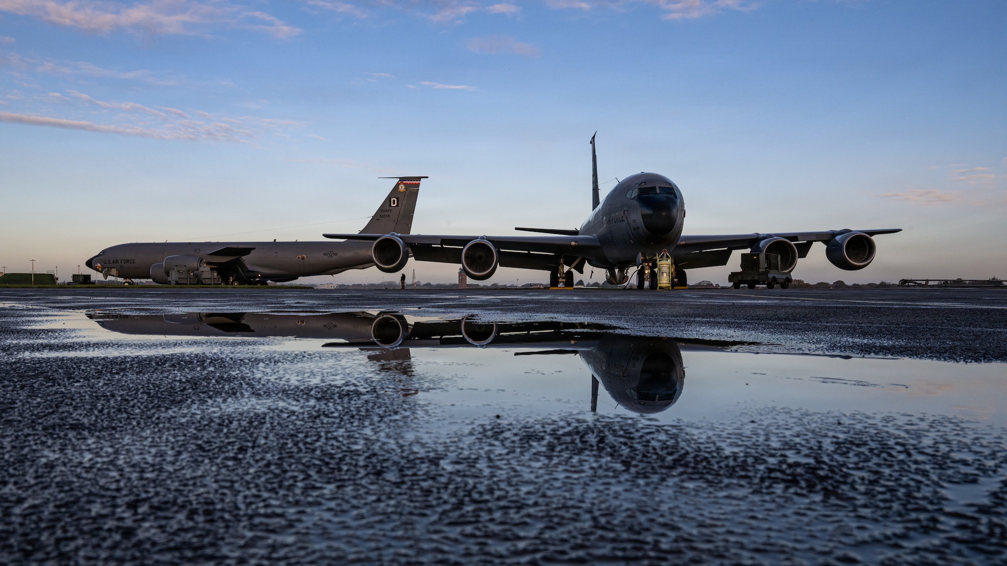 Two KC-135s sit on the flight line with their images reflected a puddle of water
