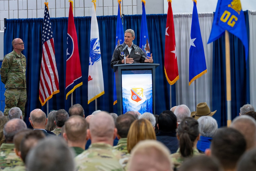 A man in a flight jacket stands at a podium addressing a crowd of military members.