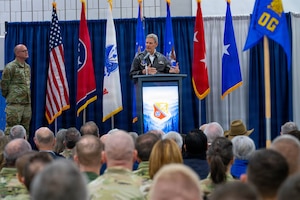 A man in a flight jacket stands at a podium addressing a crowd of military members.
