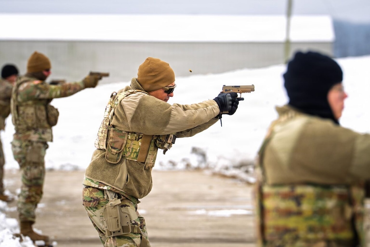 Service members wearing camouflage military uniforms practice shooting pistols outside. There is snow on the ground in the background. A shell casing is shown flying through the air.