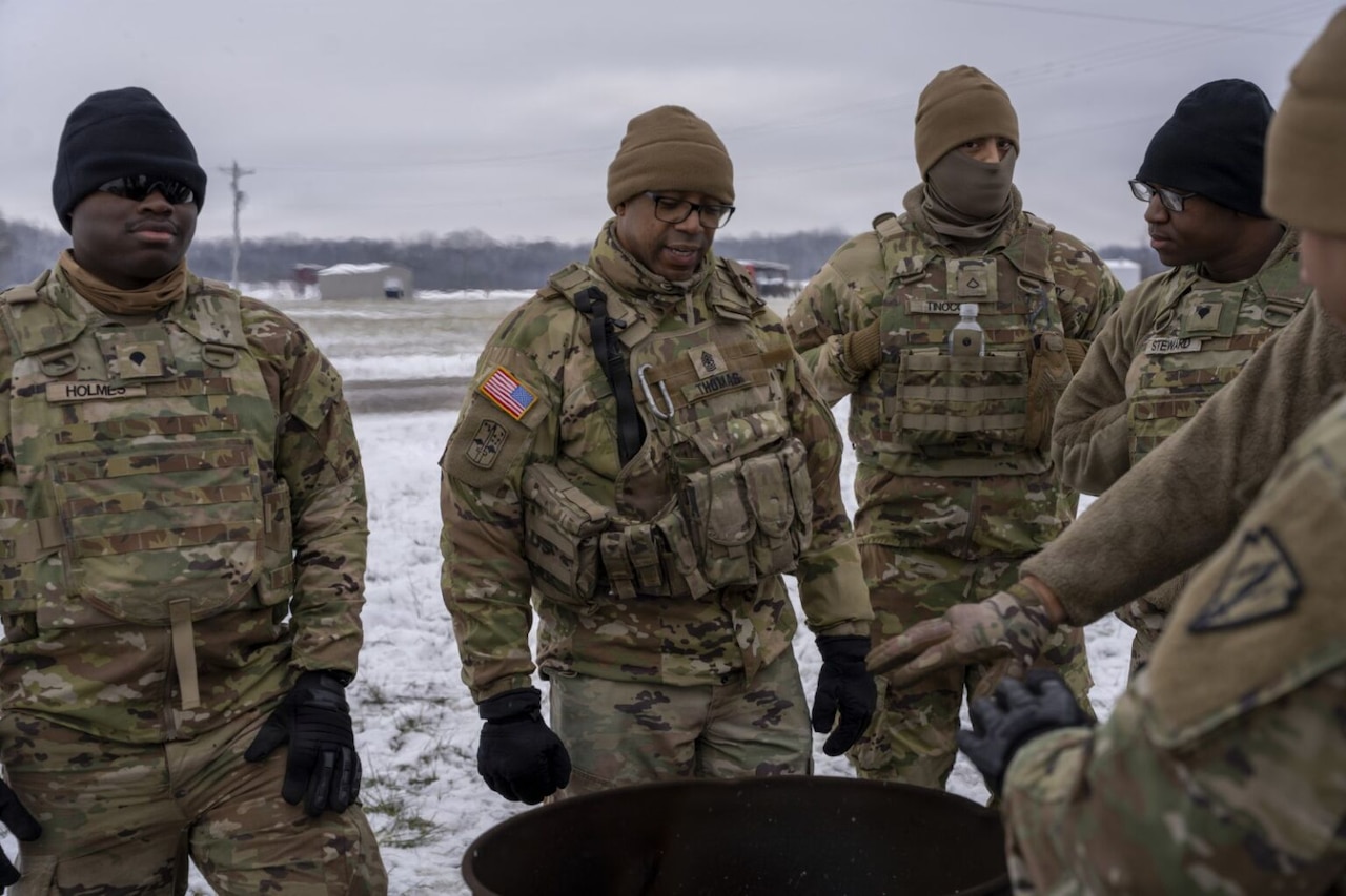 Service members wearing camouflage military uniforms, beanies and gloves stand around a barrel fire. There is snow on the ground in the background.