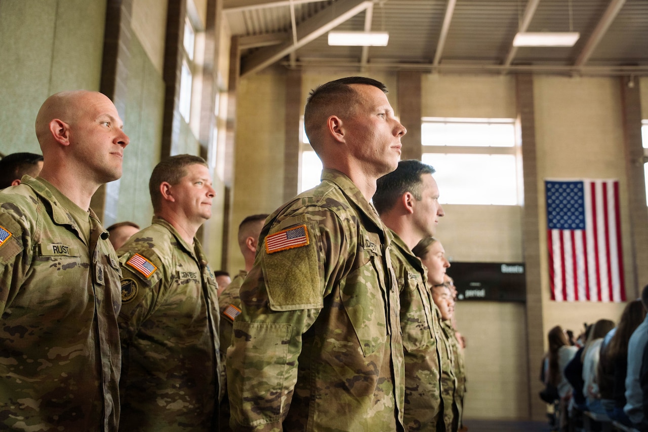 Service members wearing camouflage military uniforms stand in rows at attention during a departure ceremony. The American flag is hanging on the wall in the background.
