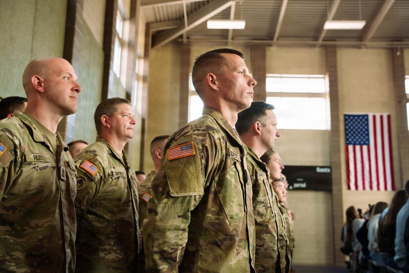 Service members wearing camouflage military uniforms stand in rows at attention during a departure ceremony. The American flag is hanging on the wall in the background.