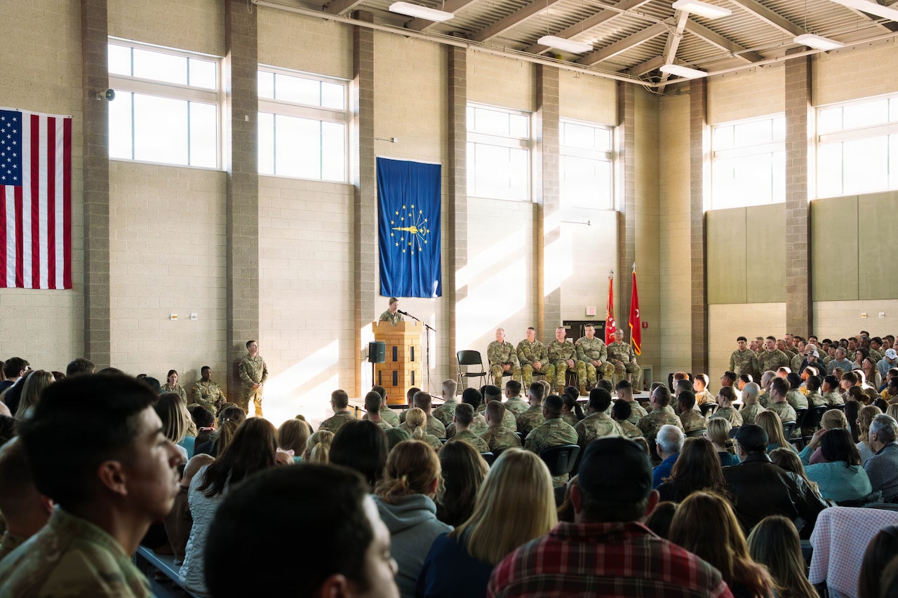 A woman wearing a camouflage military uniform stands in front of a lectern while speaking to an audience of people in camouflage military uniforms and casual attire. The people are seated in a large auditorium with the American and Indiana state flags hanging on the walls.