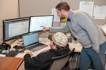 Two people are sitting at a computer looking over technical drawings.