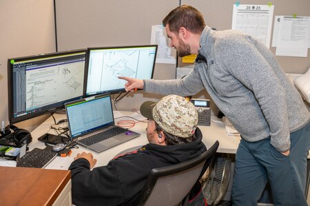 Two people are sitting at a computer looking over technical drawings.
