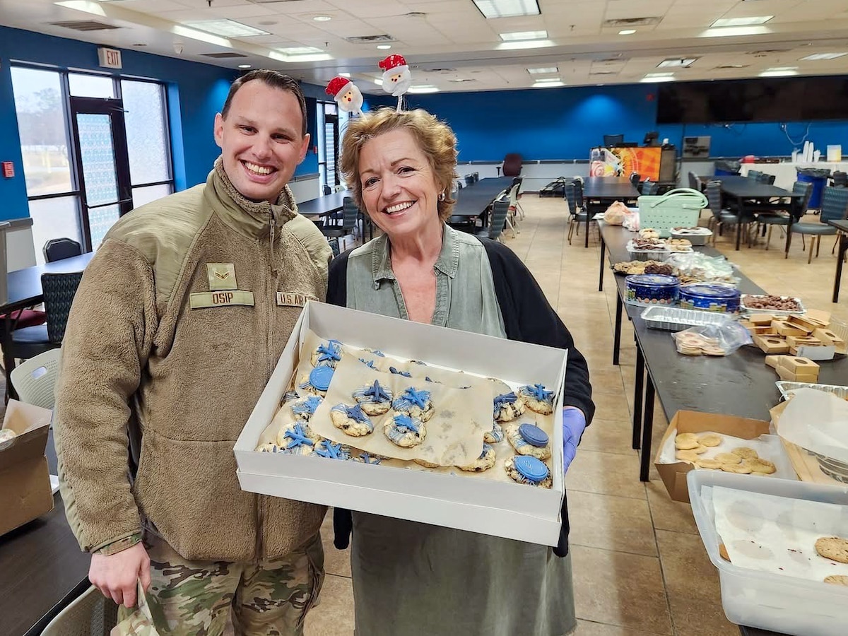 An Airman and another volunteer smile at the camera while holding a box of cookies that feature a blue design