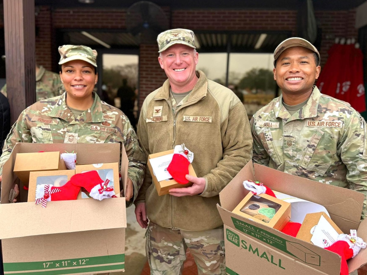 three Airmen in uniform smile at the camera while holding boxes of cookies