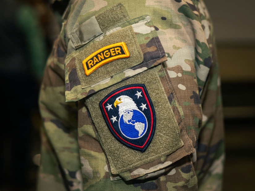 An up-close picture of the shoulder of a camouflage military uniform with a tab that says "Ranger" on top and a patch with an eagle head over a globe with four white stars depicted on a black background.