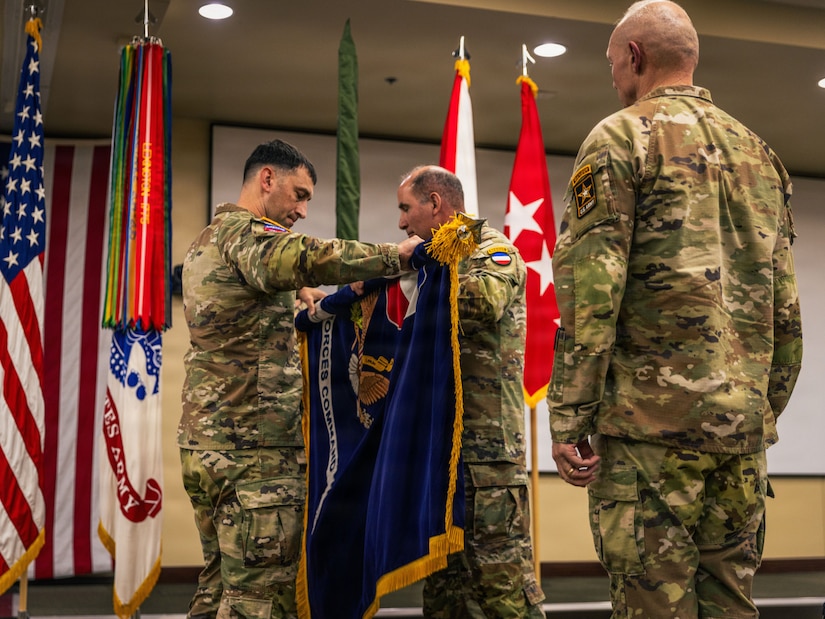 Two men wearing camouflage military uniforms roll up a dark blue flag with gold fringe as a man in similar attire watches in the foreground; the American flag and other military flags are behind them.