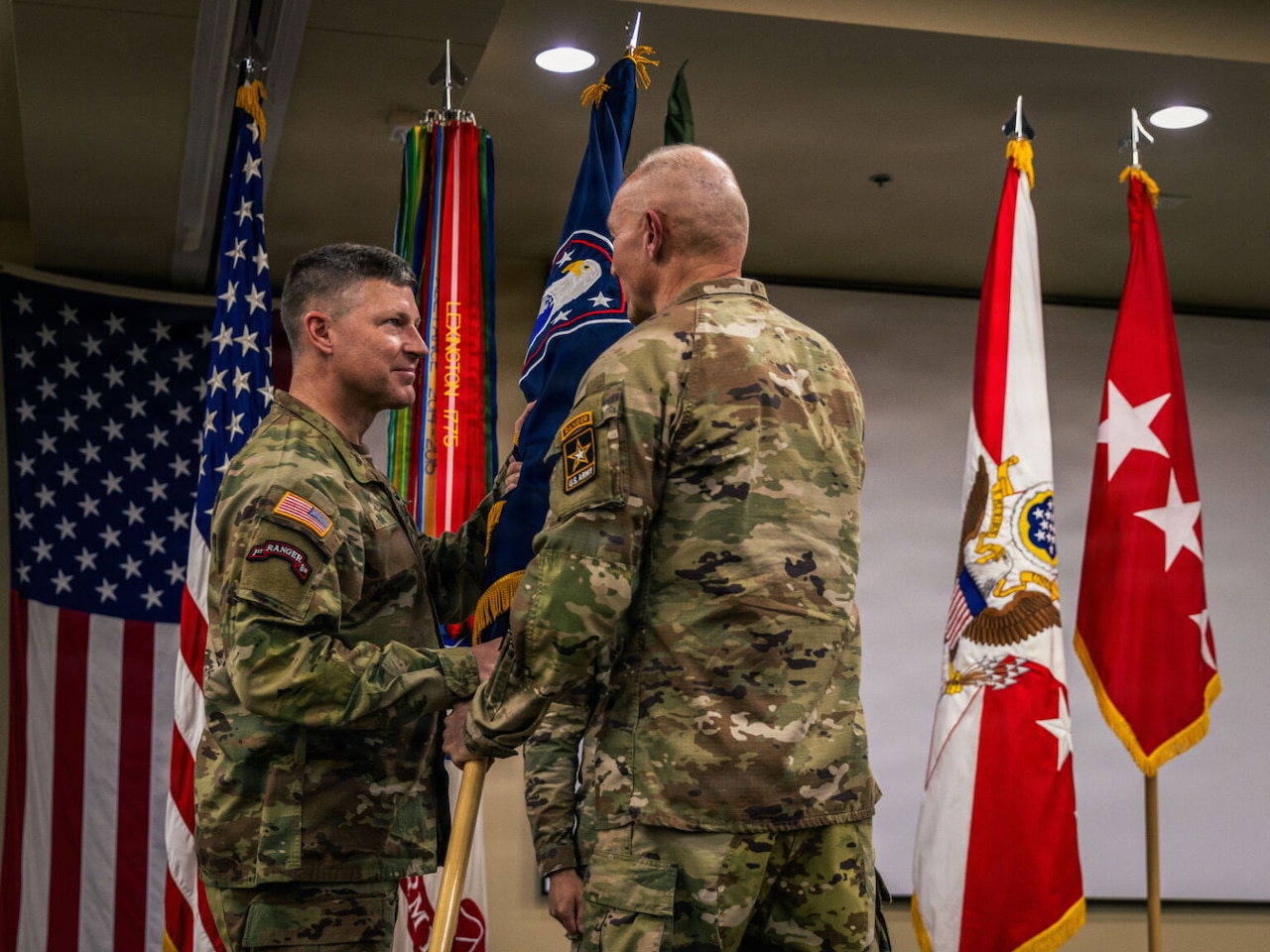 A man wearing a camouflage military uniform passes a guidon with a flag to another man in similar attire; the American flag and other military flags are behind them.