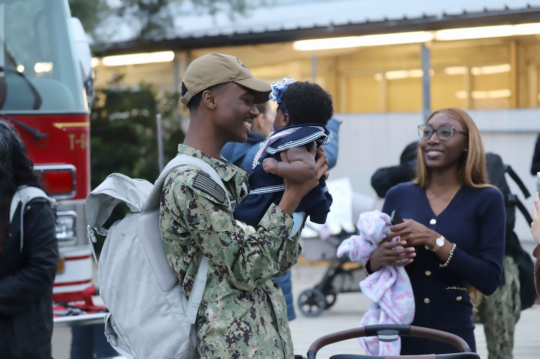 A sailor wearing a backpack smiles while holding a baby as another loved one watches from the right.