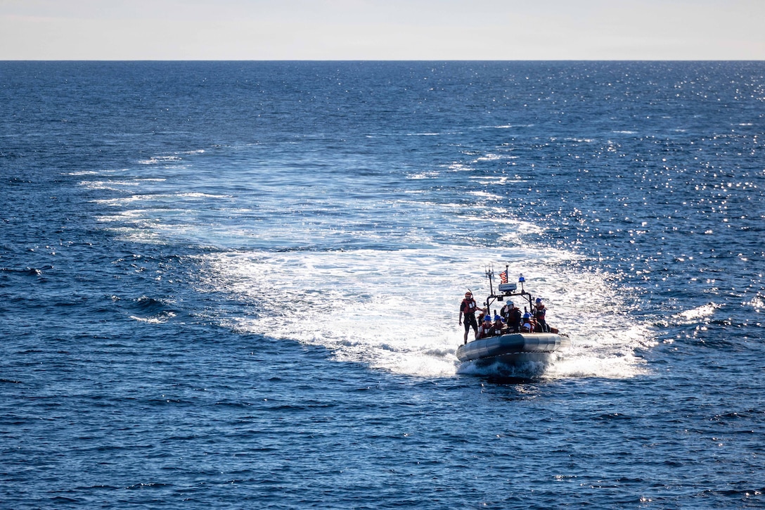 Marines and sailors wearing red life vests ride in a small boat in a body of water during the day.