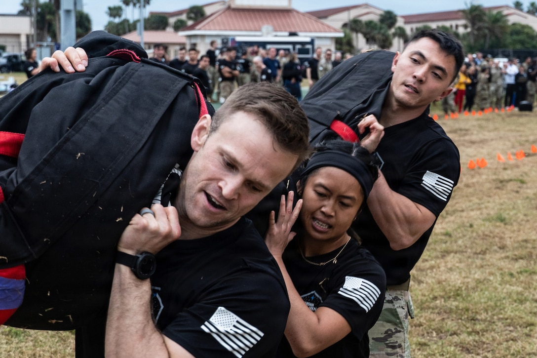 Three guardians carry a long sandbag in a field as spectators watch in the background.